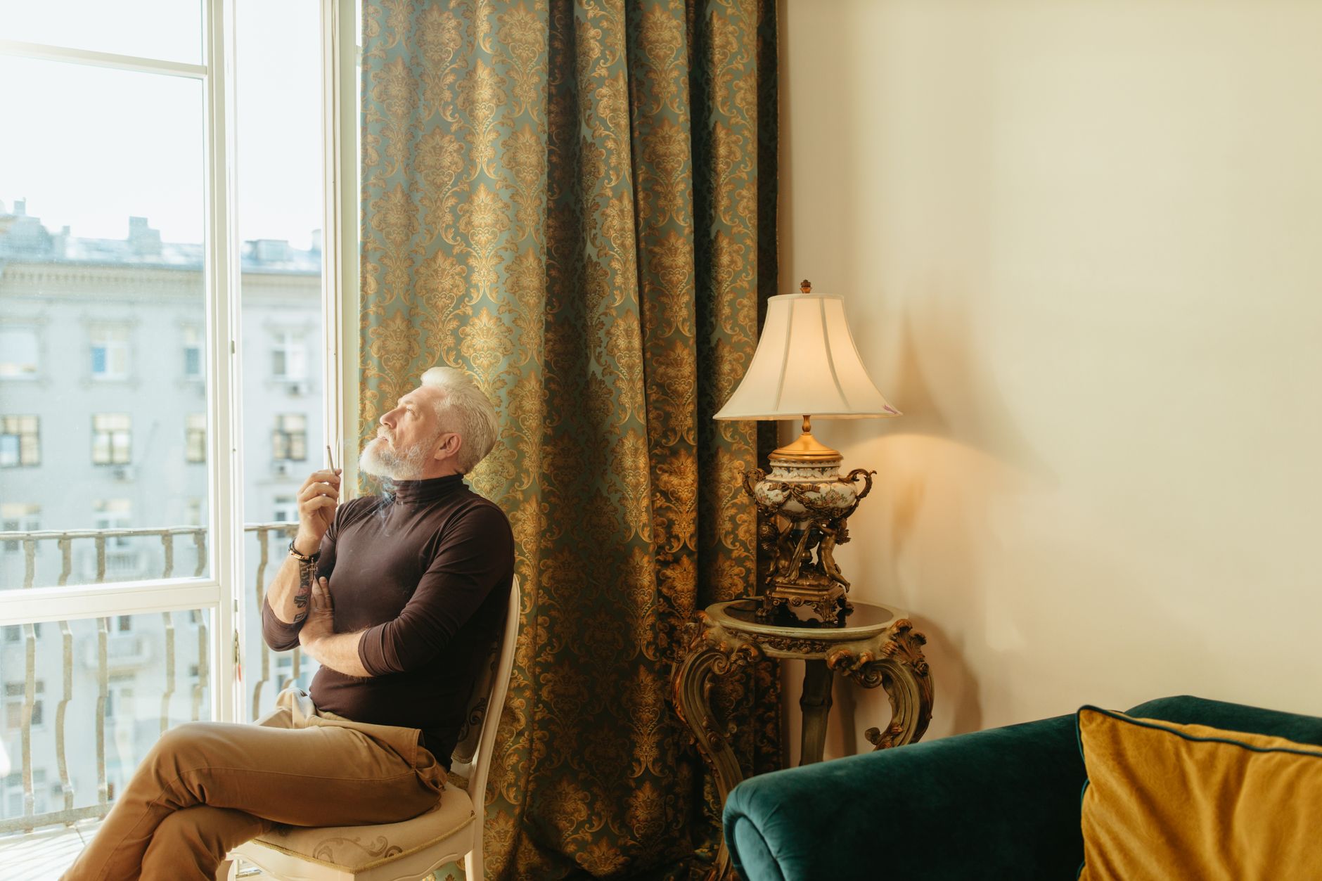 A senior man with a beard reflects while sitting indoors, elegantly framed by ornate curtains and a vintage lamp.