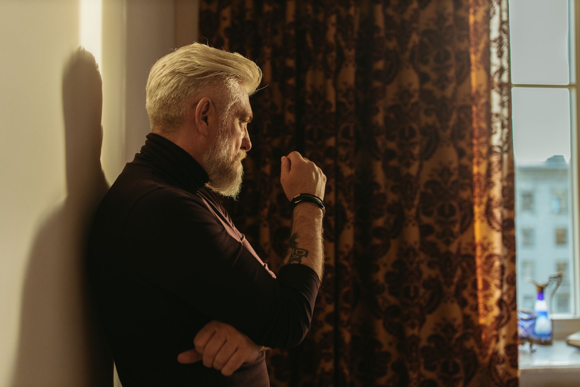 Side view of a senior man with gray hair in thought by a window with rich patterned curtains.