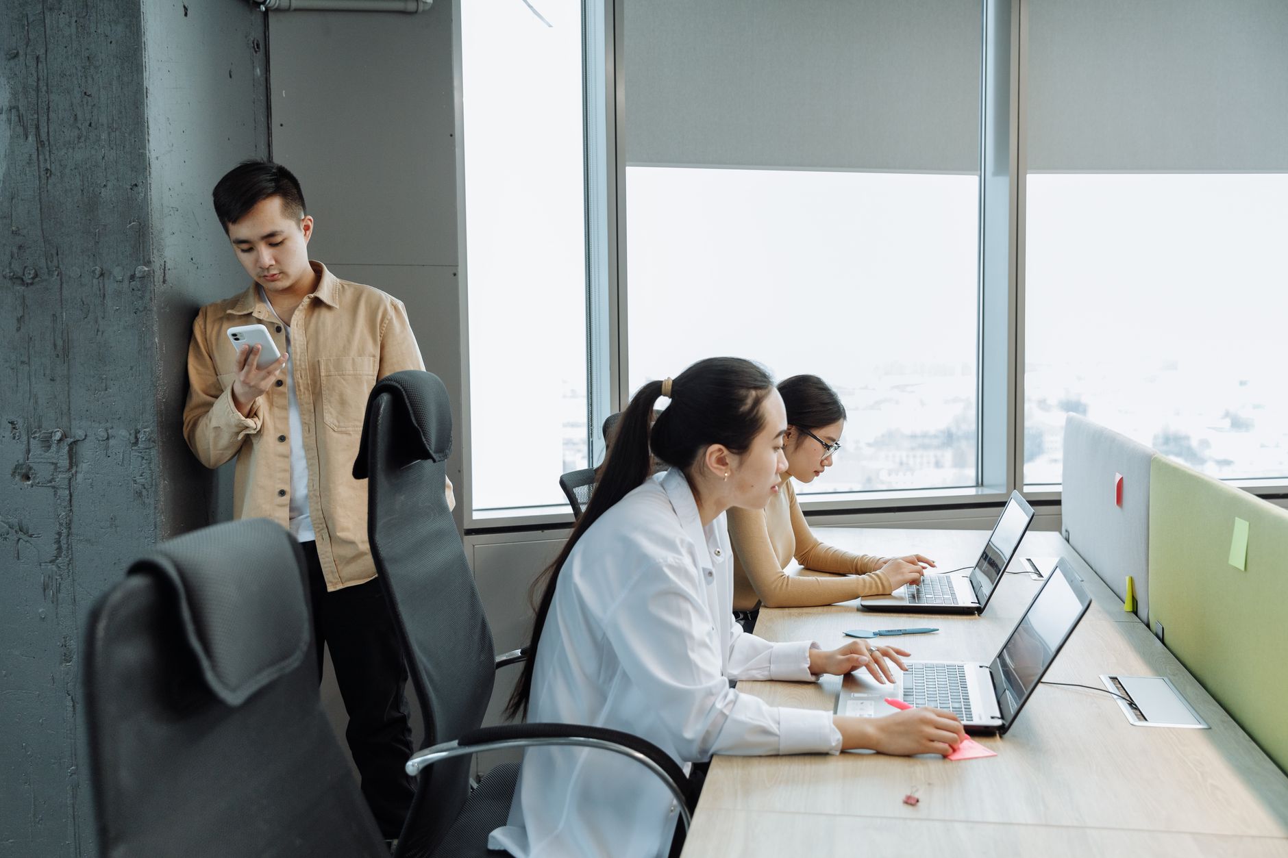 Three professionals working on laptops in a modern office setting with large windows.